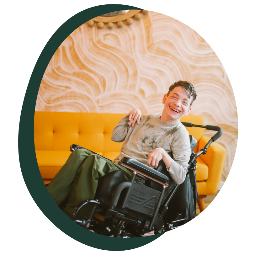 A young white man with a visible disability sits in a wheel chair smiling at the camera. He is wearing green pants and a grey sweater. He has short blonde hair. 