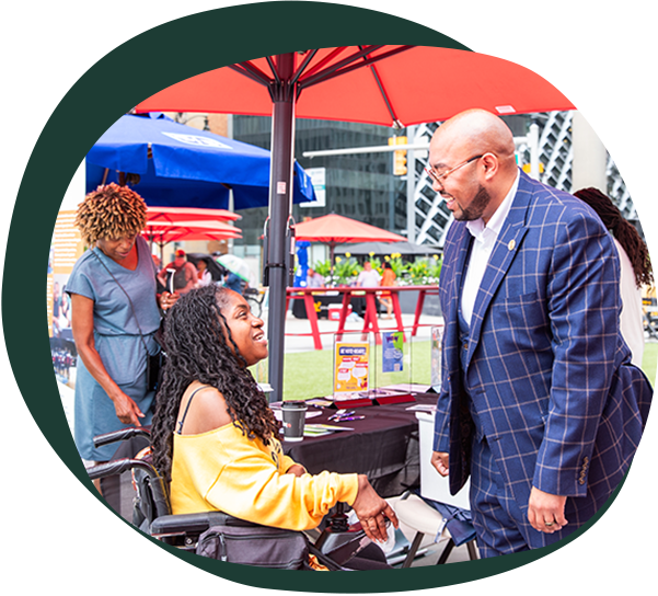A middle aged black man in a blue suit shakes hands with a younger black woman in a wheelchair. She is wearing a yellow shirt and has long braids. 