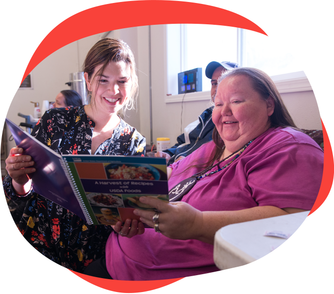 A healthcare worker (or nurse) in scrubs reads a book with an older white woman, both smiling.