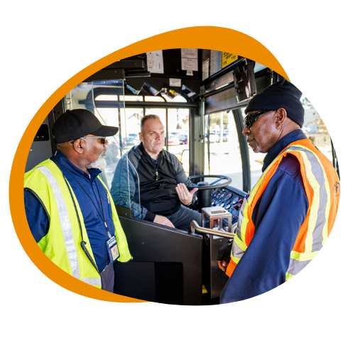 Two older black men in safety vests stand and speak to a white bus driver inside the bus next to the steering wheel. 