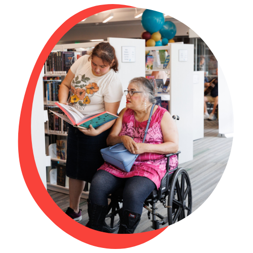 A young white woman who is standing and reading to an older white woman in a wheelchair. They in between library stacks.
