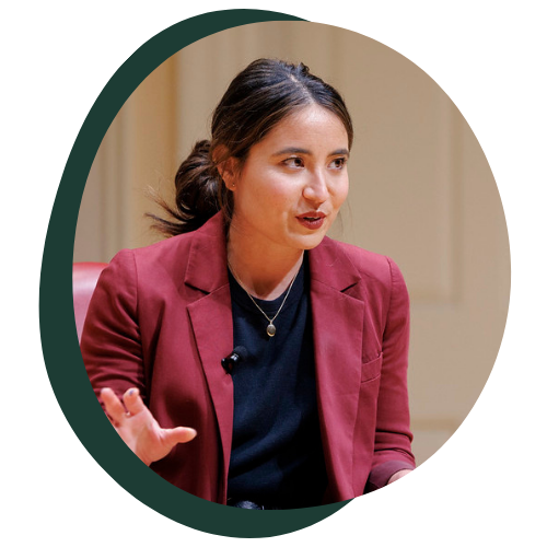 A young Asian woman in a maroon suit jacket and black shirt speaks to a crowd. 