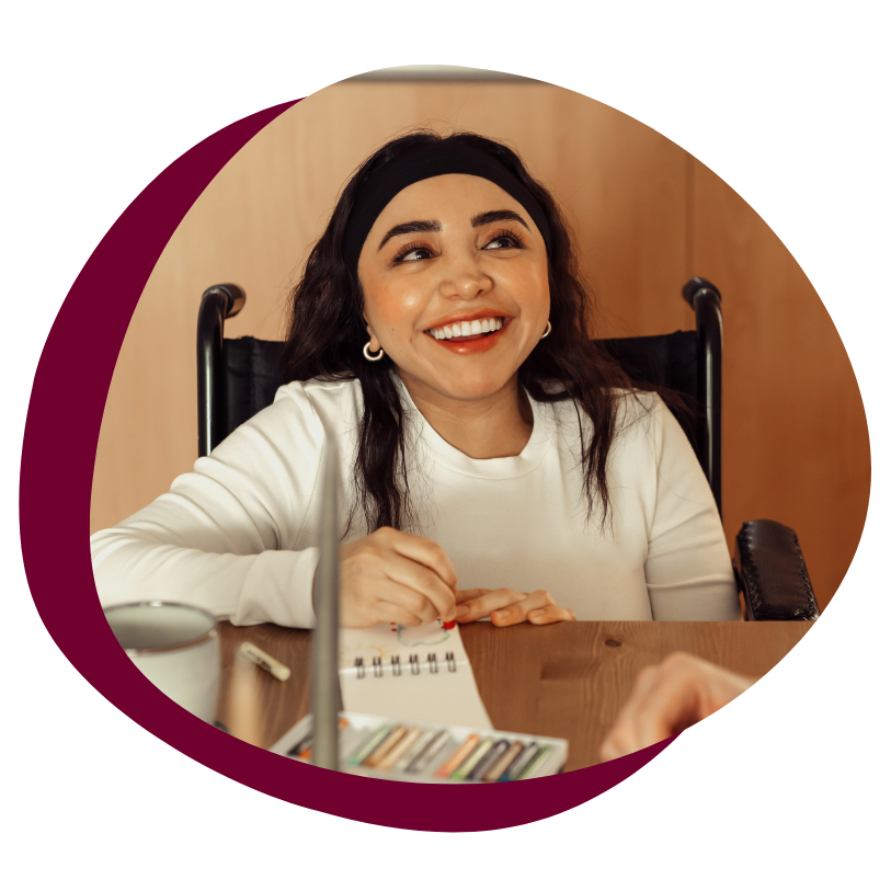 A young latina woman sits in her wheel chair at a table smiling and writing on a notepad. She has long black hair held back with a black headband. She is wearing red lipstick with silver hoop earrings and has a white long sleeve shirt on. 