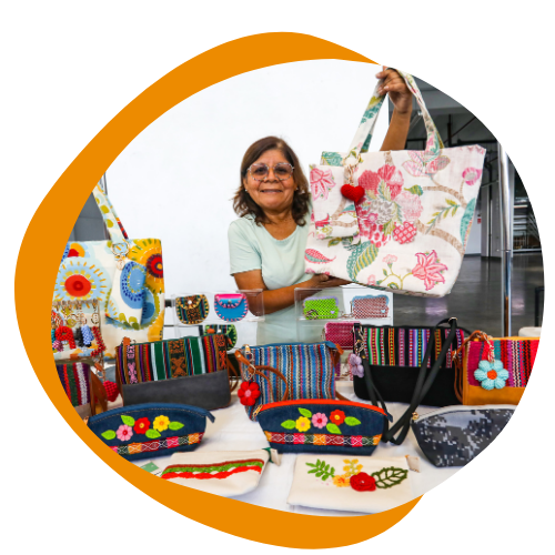 An older woman of color is holding up a colorful purse she is selling. In front of her is a table of other colorful purses she is selling. She is wearing glasses. Has short straight greying hair and a teal short sleeve shirt. 