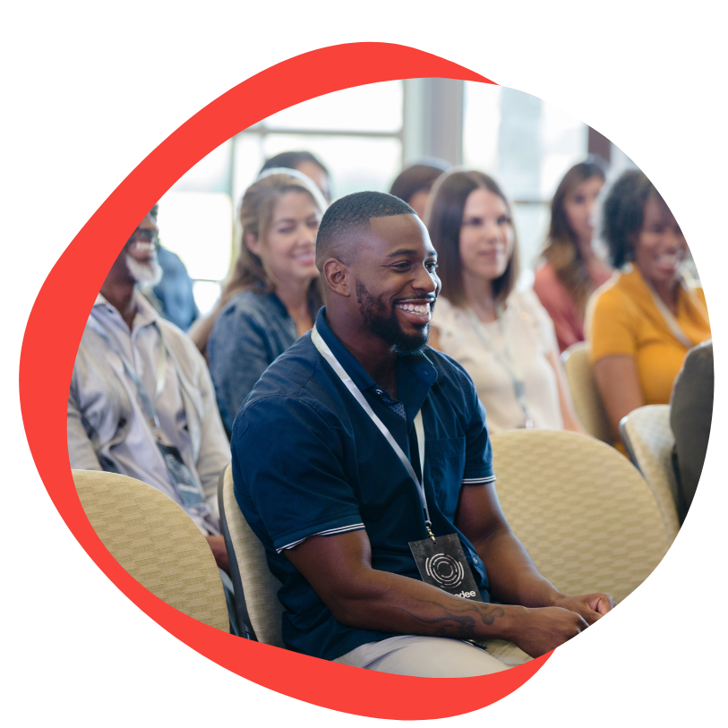 A young black man sits on a tan chair in a conference setting. He has a buzz cut with a short beard. There are people sitting in the same chairs in rows behind him. He is wearing a short sleeve blue collared shirt and wears a lanyard and name tag around his neck. He is smiling.