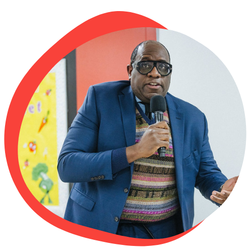 An older black gentleman stands and speaks into a microphone. He is wearing large black glasses, a blue blazer, and a colorful sweater.