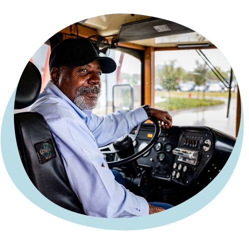 An older black man with a blue collard shirt black hat and beard sits in the drivers seat of a bus. He is turning behind to look at the camera. 