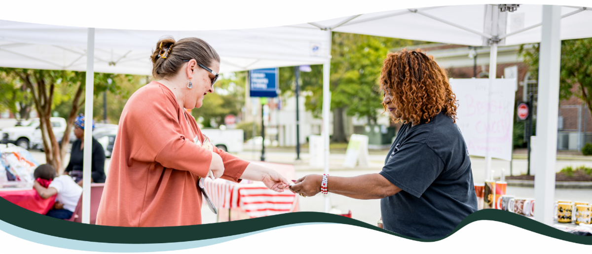 A white woman hands payment to a Black woman vendor at a farmers market stall, exchanging goods with a smile.