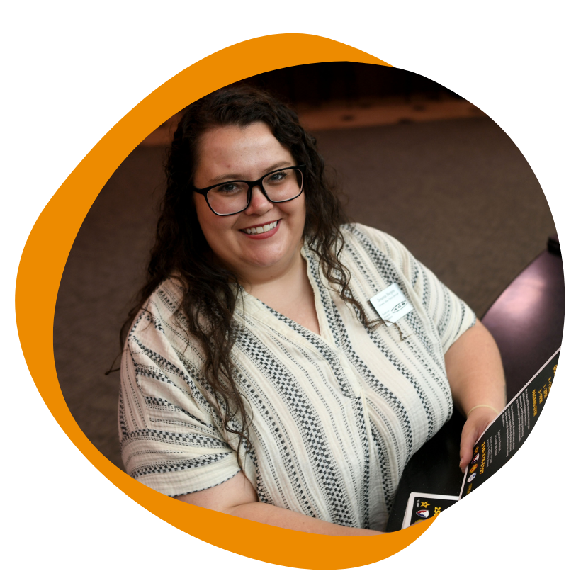 A white woman with long dark hair sits at a table. She is wearing glasses and a beige patterned short sleeve shirt. She is also wearing a name tag and smiles at the camera. 