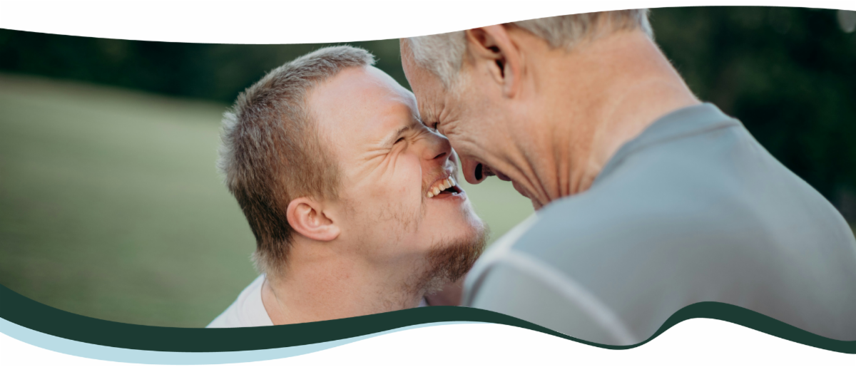 An older white man smiles and touches foreheads with a younger white man with down syndrome.