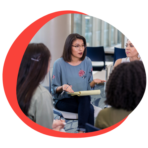 A Native American woman sits in a circle while talking and presenting to others in the room..