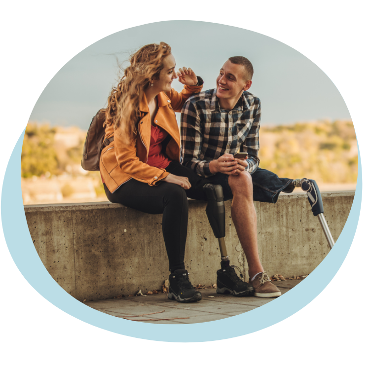 A white woman sits next to a white man with a prosthetic leg. She is wearing a tan leather jacket and has long wavy blond hair. He is wearing a flannel shirt and shorts and has black hair in a buzzcut. They sit on a concrete retaining wall in front of a sandy beach.