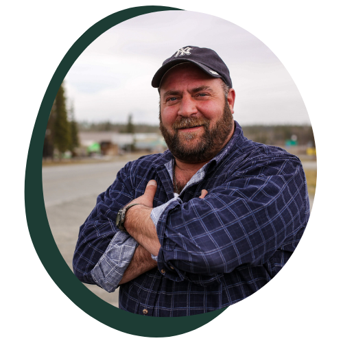 A middle aged rural white man poses for the camera. He is wearing a cap, has a medium sized beard, has his arms crossed, and wears a blue flannel. 