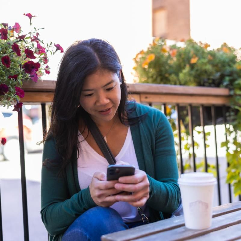 A woman looking at the phone, sitting on a patio with a coffee cup on the table.
