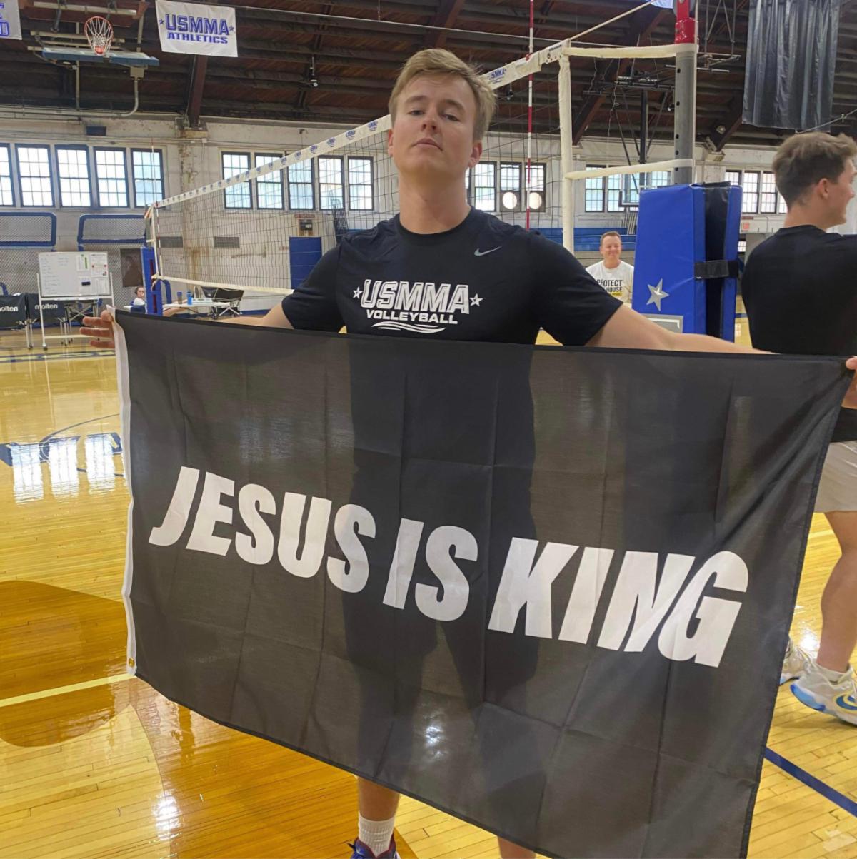 Midshipman Jackson Tolle in gym wearing USMMA volleyball shirt holding large Jesus is king flag outstretched in front of him 