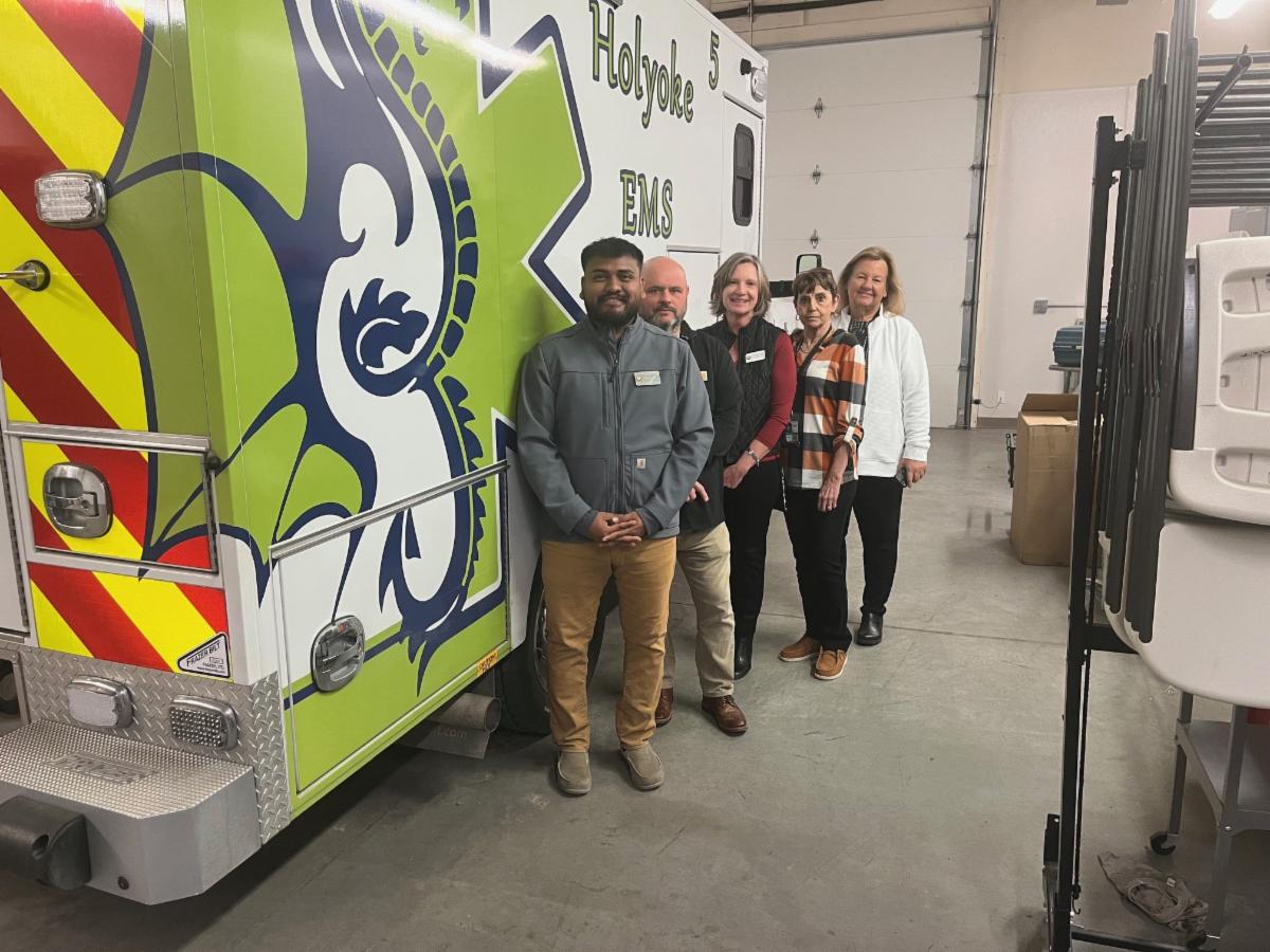 Five adults stand smiling beside a bright green “Holyoke EMS” ambulance inside a garage-like emergency services facility.
