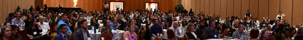 Group of 500+ individuals sitting in a large room surrounding circular tables. The audience is listening to a presenter on stage.