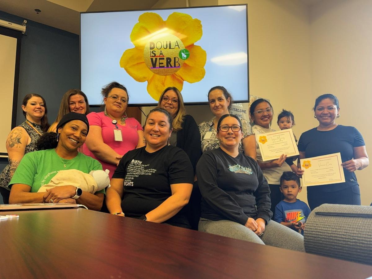 Group of women sitting at a board room table, smiling, in front of a TV screen that reads "Doula is a verb" on a sunflower design. Two women are the right are holding certificates. Several women are holding infants in the photo.