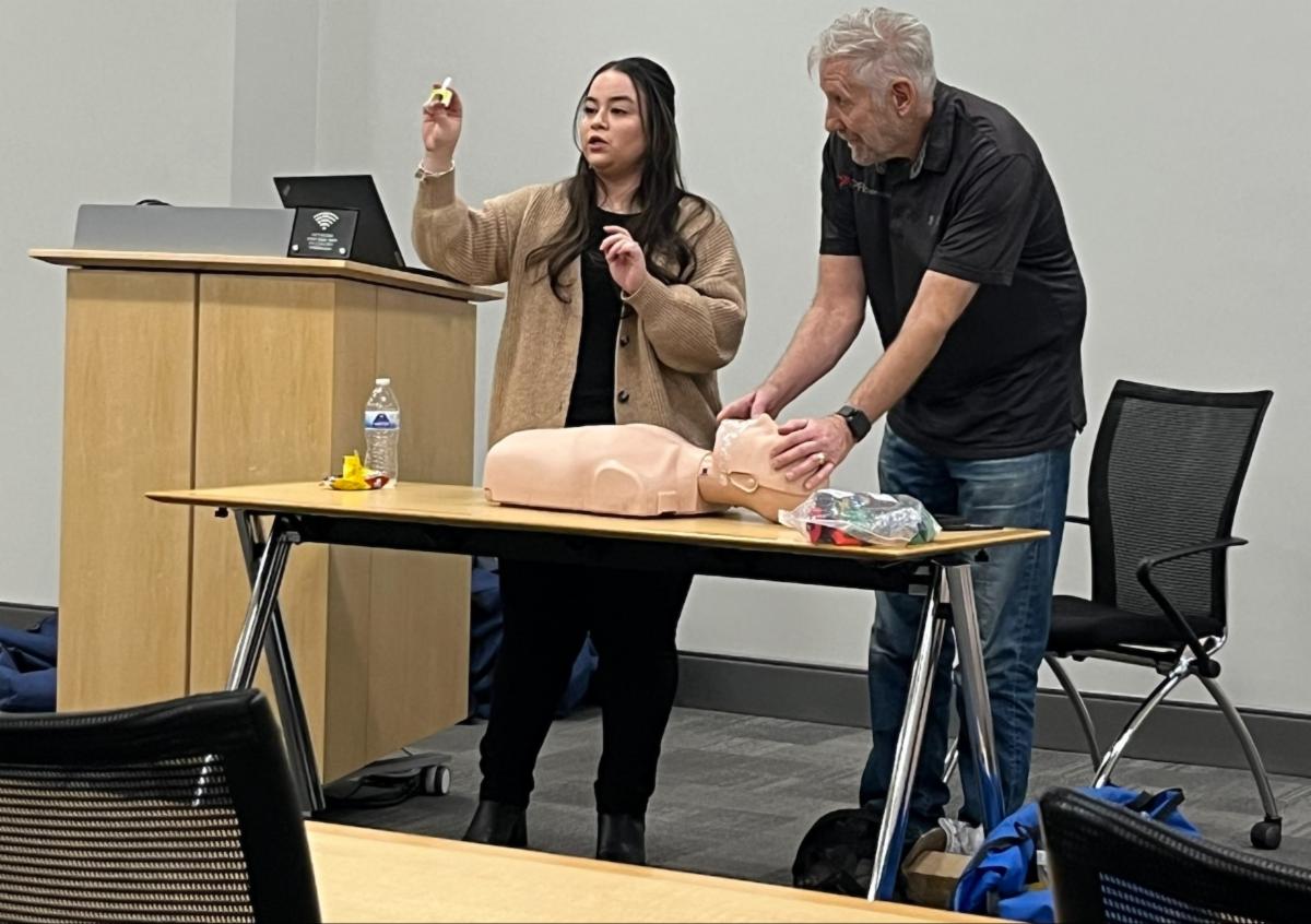 Two people demonstrate emergency response techniques on a CPR training mannequin, with one holding a small device while the other positions the mannequin on a table in a classroom setting.