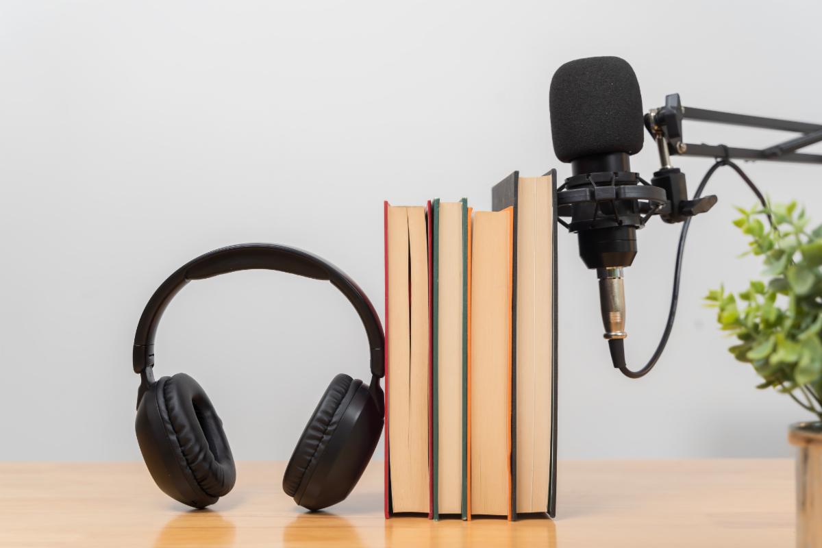 Close up of stacked books with headphones and a professional microphone on a wooden desk symbolizing podcast audiobook education online learning knowledge sharing reading digital communication.