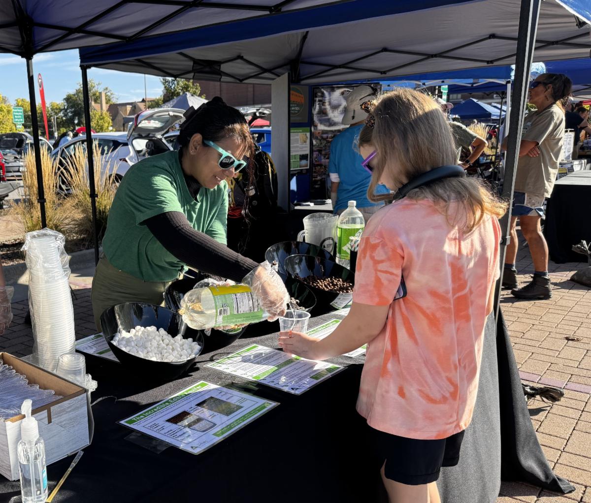 Photograph shows an outdoor community event booth where a volunteer in sunglasses serves shaved ice or a cold drink to a child. Black canopy tents, colorful informational flyers, cups, ice containers, beverage bottles, and other attendees create a busy outreach setting.