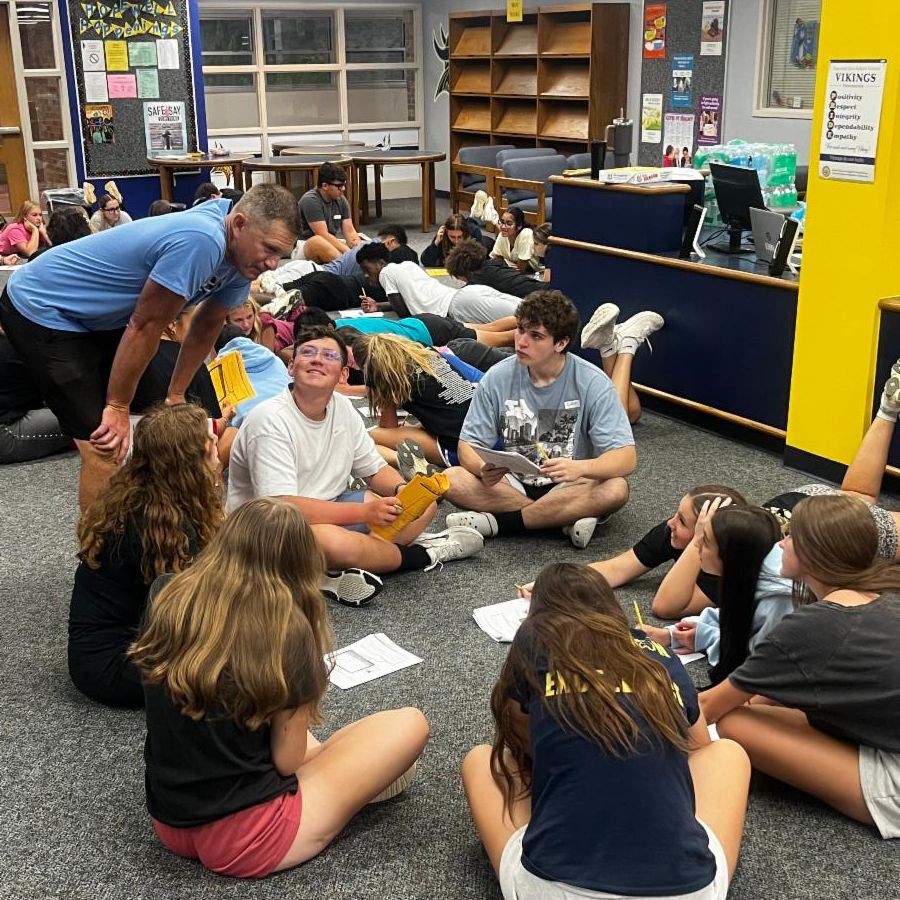 Students siting in circle on floor with teacher standing next to them
