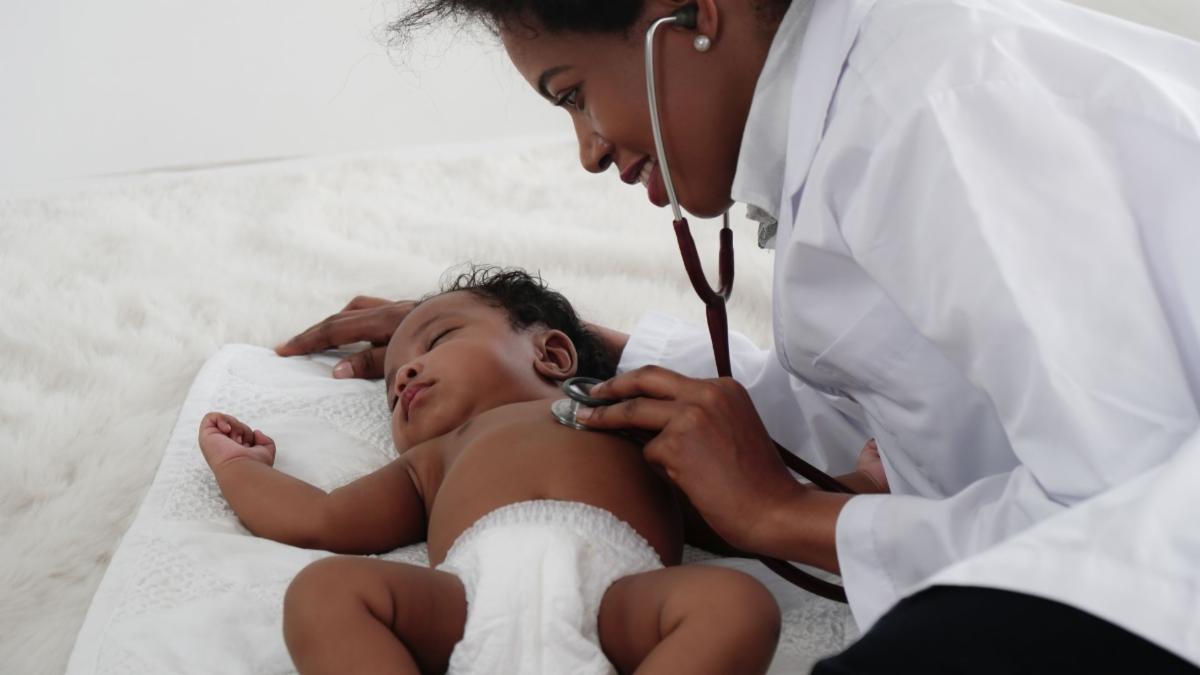A medical professional in a white coat uses a stethoscope to examine a baby lying on a white blanket.