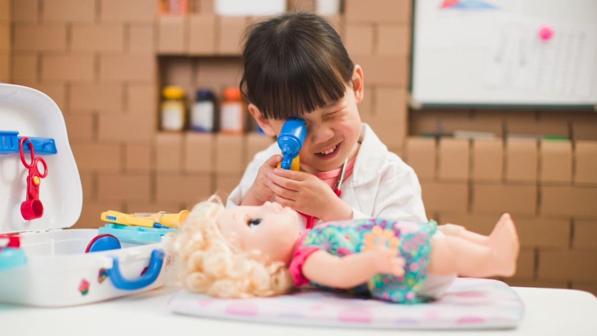 A child dressed as a doctor examines a doll with a toy stethoscope, surrounded by a toy medical kit.
