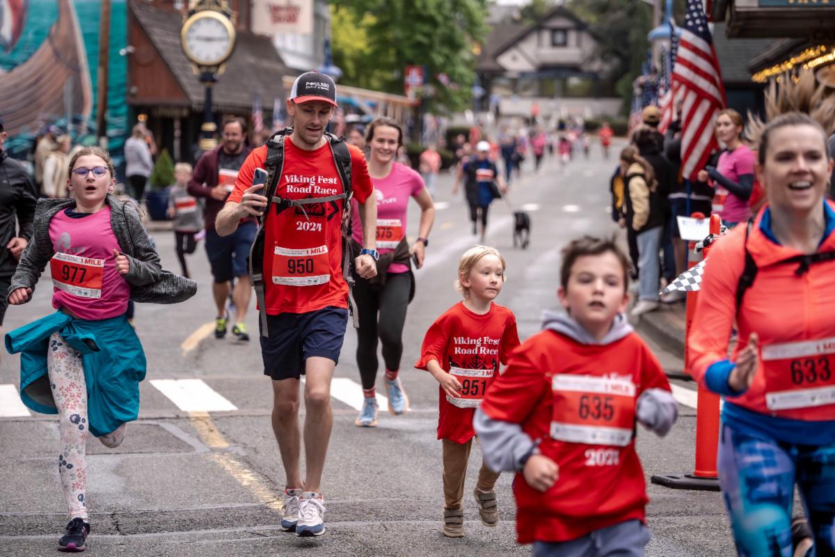 People running down historic downtown Poulsbo Front Street wearing orange and hot pink t-shirts with runner numbers on their shirts in the Viking Fest Road Race.