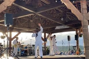 6 people onstage at the Waterfront Park in their white Navy attire singing and playing instruments in front of an audience.