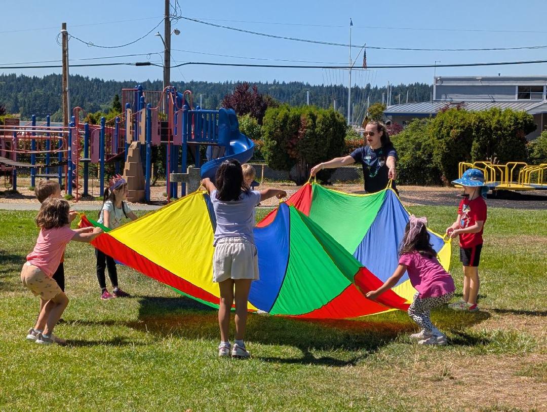 A group of children with an adult playing with a colorful parachute on a playground.