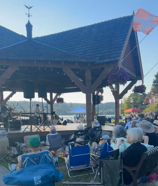 A band onstage playing to lots of people sitting on beach chairs on the grass and sidewalks. The Norwegian flag is waving in the wind.