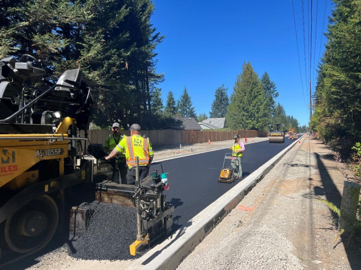Workers in yellow vests and hats working on paving the road with big paving equipment.