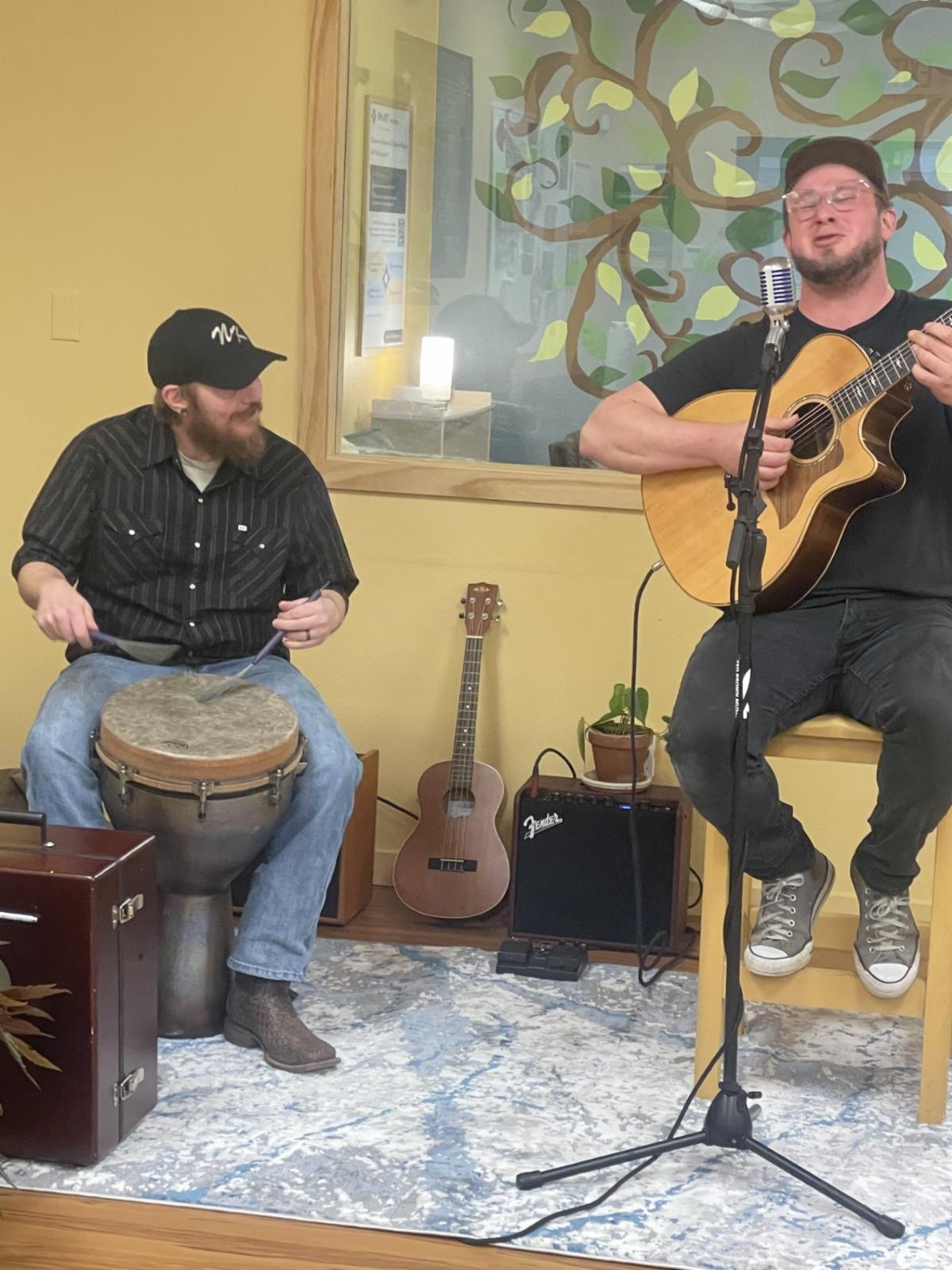 Man with cap playing drum and man with cap playing guitar and singing at Recovery Cafe.