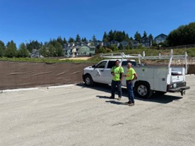 Two men in yellow shirts standing by a white work truck on the new gravel parking area. Dirt and vegetation and houses and blue sky is behind them. 