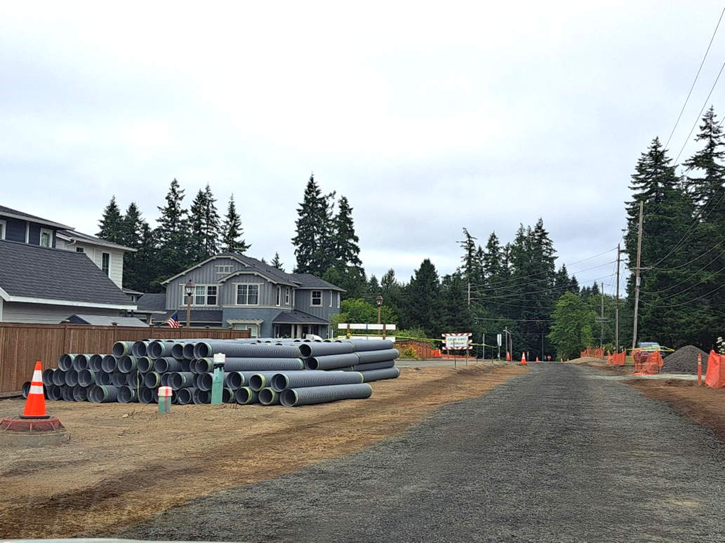 A gravel road with rows of large gray pipes next to a fence. Orange caution cones and orange fencing in the work zone.