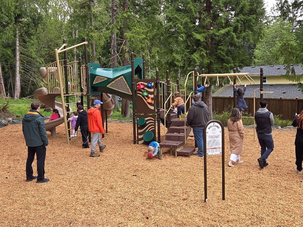 Adults watching children playing on the playground equipment at the Rotary Morrow Park. The ground is covered in fresh wood chips and tall green trees are in the background.