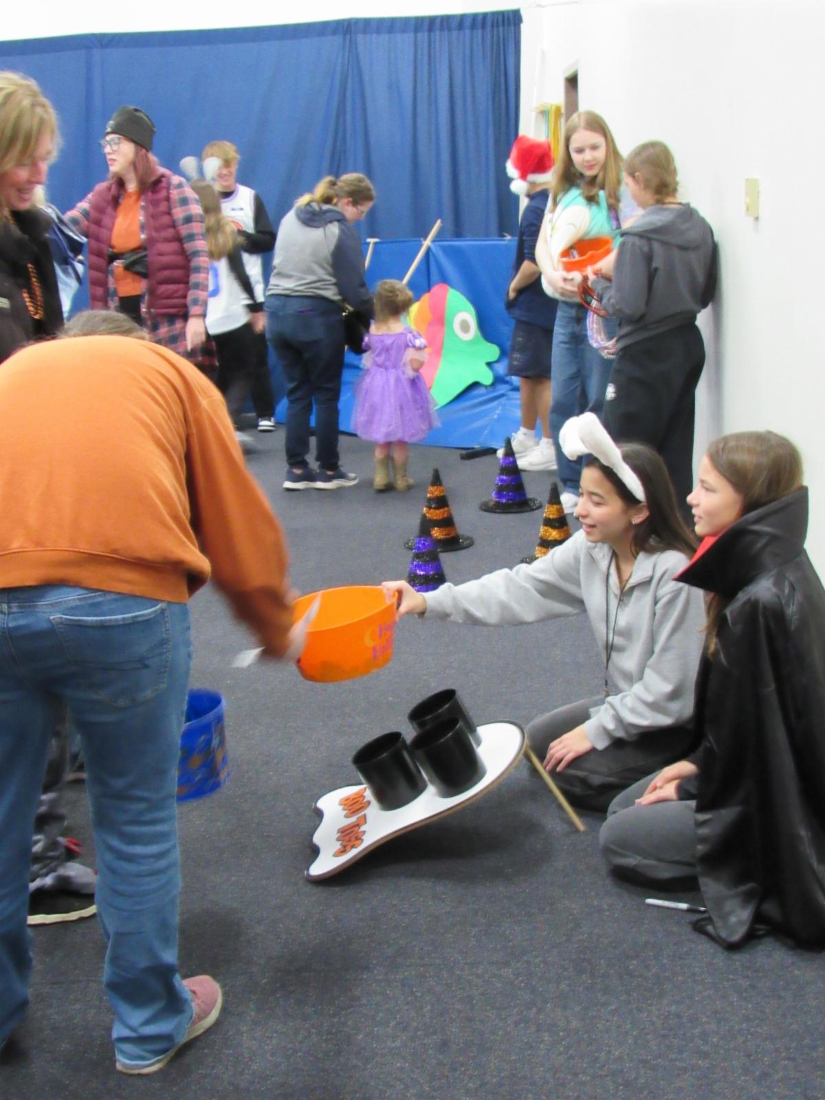 Children with their parents playing games at a Halloween carnival. Older kids are running the games. 