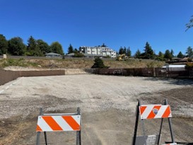 2 caution signs in front of the new gravel parking area. A hill with dirt and vegetation and a house is behind the parking area.