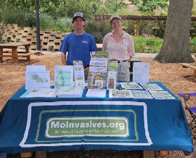 Clifford Barratt and Lilly Germeroth, both MPF staff members, standing behind a picnic table used as a booth table, with the MoIP table covering over it and many free handouts on invasive and native plants. 
