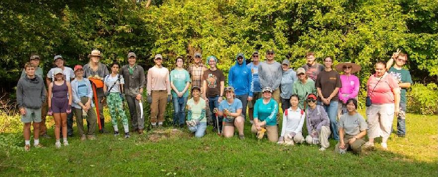 About 20 volunteers standing in two rows at the Missouri Conservation Corps volunteer event at Again Park in Columbia on August 8. They are standing on mowed grass with threes behind them.