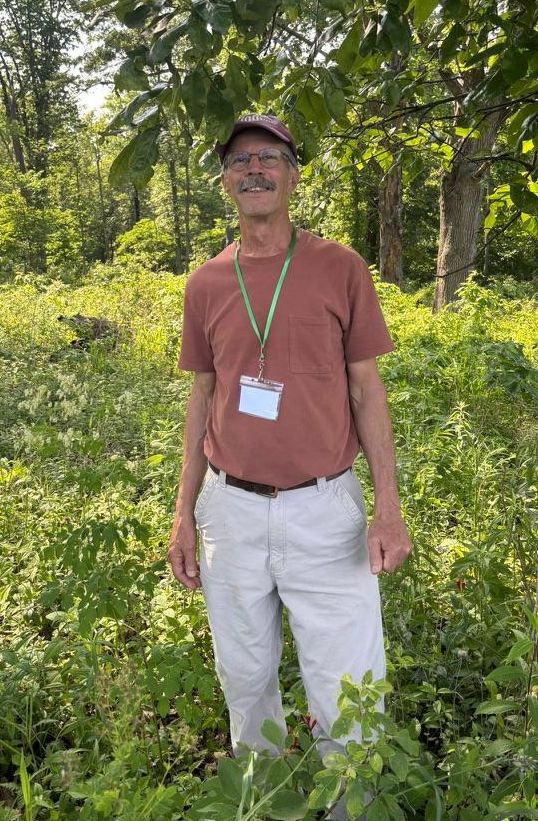 MoIP 2026 Awardee Mike Smith standing in a woodland cleared of invasvies. There is dappled sunlihgt and a tree branch above his head. He has a cap, a clay colored t-shirt, a meeting lanyard around his nectk and light colored pants on.