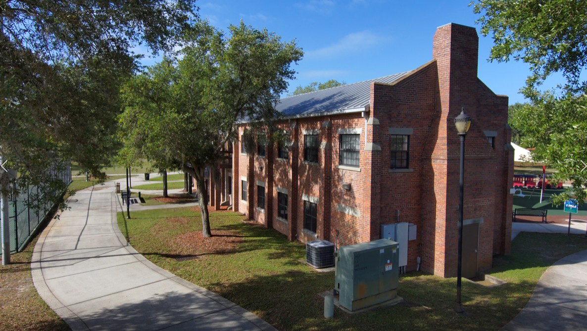 A twoâstory brick armory building with a metal roof, tall rectangular windows and a prominent chimneyâlike tower on one end. The structure sits beside a walkway with trees and open green space.