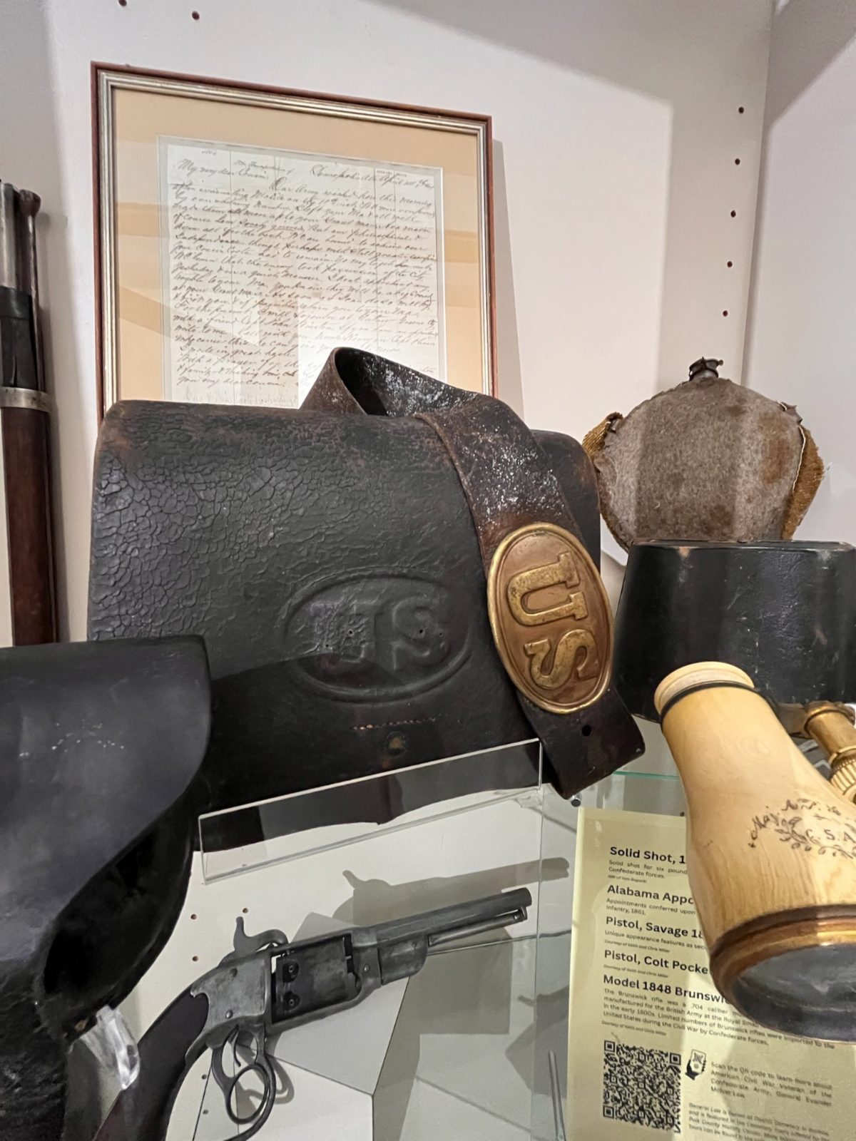 Museum display case featuring Civil War–era artifacts, including a black leather “US” cartridge box, a matching belt with a brass “US” buckle, a framed handwritten letter, a worn cloth-covered canteen, a revolver, and a wooden-handled powder flask, arranged with a yellow identification card listing the items.