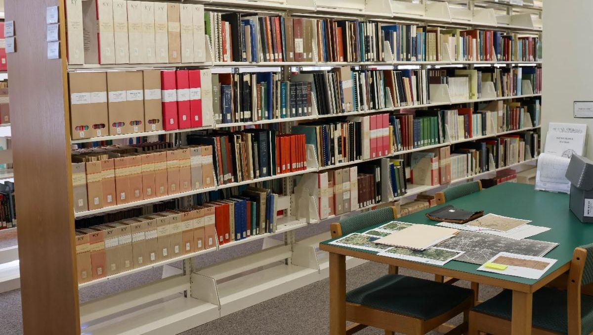 A genealogical research room with shelves of labeled books and archival boxes organized by state. A green table in the foreground holds open folders, maps, and papers, with chairs arranged for researchers.