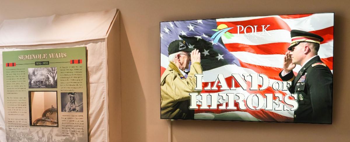 Exhibit area featuring a Seminole Wars informational panel with images and text, next to a digital display showing two service members saluting in front of an American flag with the title Polk County Land of Heroes.