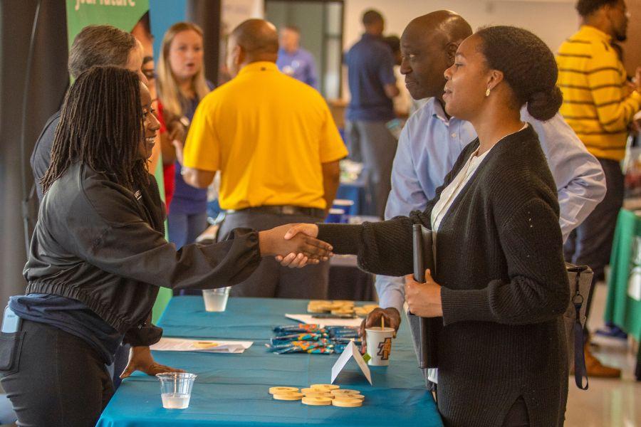 The caption reads: A student makes a connection with one of the recruiters during Ag Awareness Day at the University Farm Pavilion. The career fair gave students a chance to network and job hunt. The image displays a young woman in a black sweater shaking hands with an older man in a blue shirt. The woman has long black hair and is smiling. The man has a serious expression on his face. They are standing at a table with a blue tablecloth, surrounded by other people. There are several cups and some snacks on the table. 