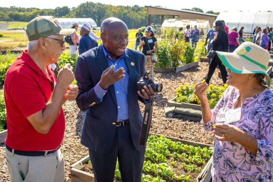 The caption reads: Farm safety expert Jimo Ibrahim, Ph.D., chats with two of the attendees at&nbsp;Cooperative Extension’s first Regional Small Farms Conference. Experts and growers from Virginia, North Carolina and South Carolina came together to create, and attend, the conference.The image shows three people stand in a garden setting, engaged in conversation. The man on the left, wearing a red shirt and khaki pants, listens intently as the man in the middle, dressed in a blue blazer and blue shirt, speaks while holding a camera. The woman on the right, wearing a pink floral blouse and a wide-brimmed hat, examines a small yellow flower. The scene is a mix of casual and formal, with the men's attire suggesting a professional setting, while the woman's outfit is more relaxed. The background features a bustling garden filled with various plants and trees, suggesting a lively outdoor event or gathering. The overall ambiance is one of casual conversation amidst a vibrant environment.