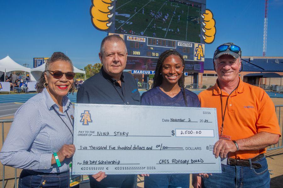 Interim Dean Shirley Hymon-Parker, of the College of Agriculture and Environmental Sciences, joins Grant Ozipko and Carroll Moseley of Syngenta US in awarding sophomore Nina Story with the 2024 Ag Day Scholarship. Story is a track and field athlete and an animal sciences major. The image shows a group of four people standing outside a stadium. Two women and two men are holding a large check. The check is made out to Nina Story for $2,500.00 for the Ag Day Scholarship. The check is dated November 2, 2024. The people are smiling and appear to be happy. The stadium scoreboard in the background shows a football game in progress. The image captures a moment of celebration and achievement.