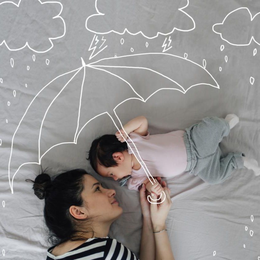 Woman and infant laying down on a bed with grey sheets with an illustration of an umbrella protecting them from rain